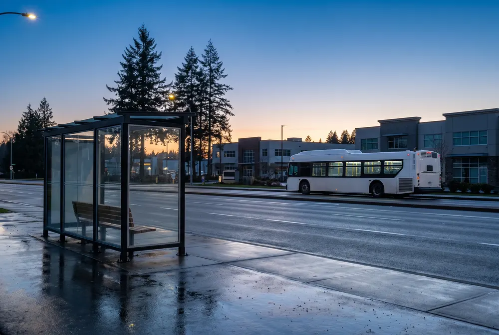 Generated scene: a glass bus shelter on a wet highway sidewalk at dusk, a plain white city bus pulling away past low modern buildings and tall evergreens.